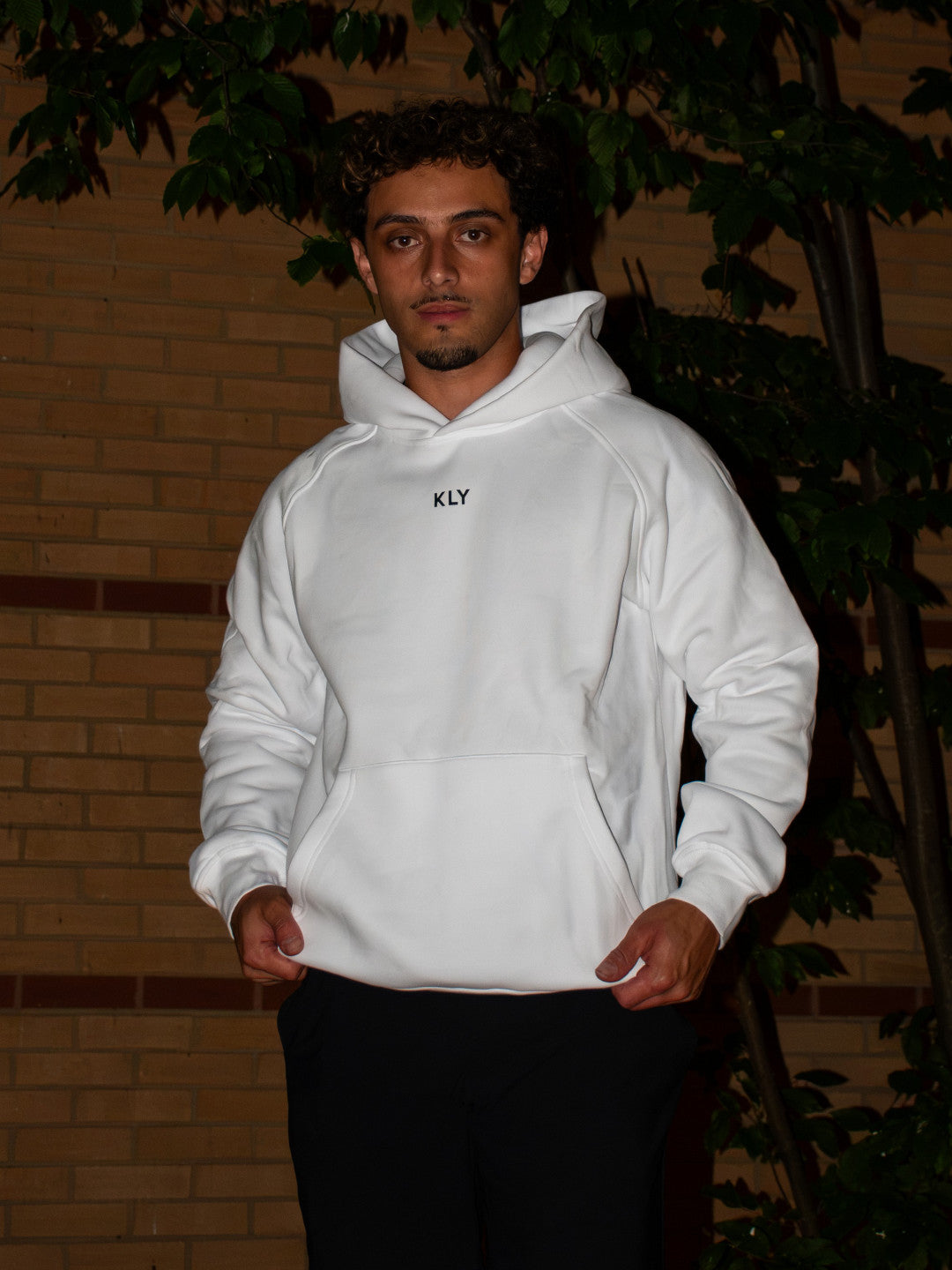 Antonio facing the camera in the KLY Original Hoodie in white at night, tree and brick wall behind him